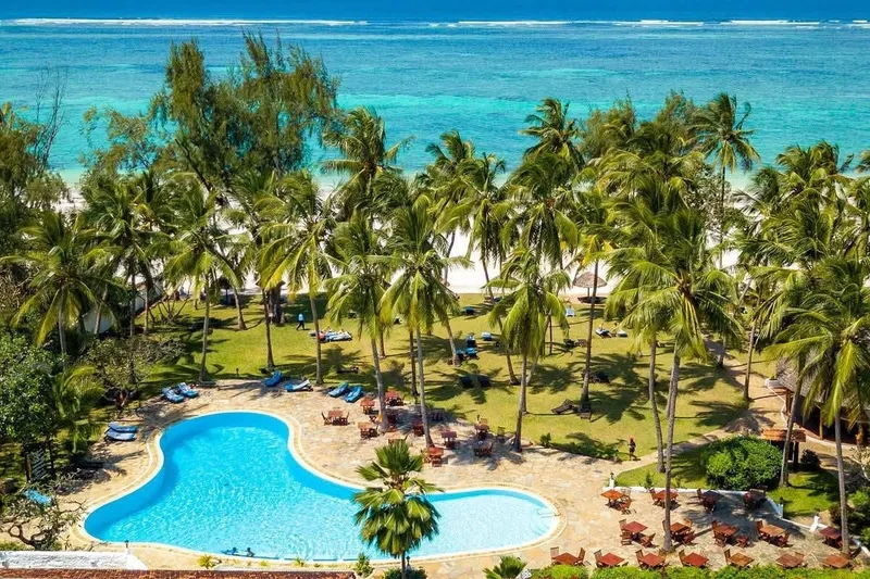An aerial view of a winding swimming pool nested within a dense grove of tropical palms near the sparkling waters of the Indian ocean.